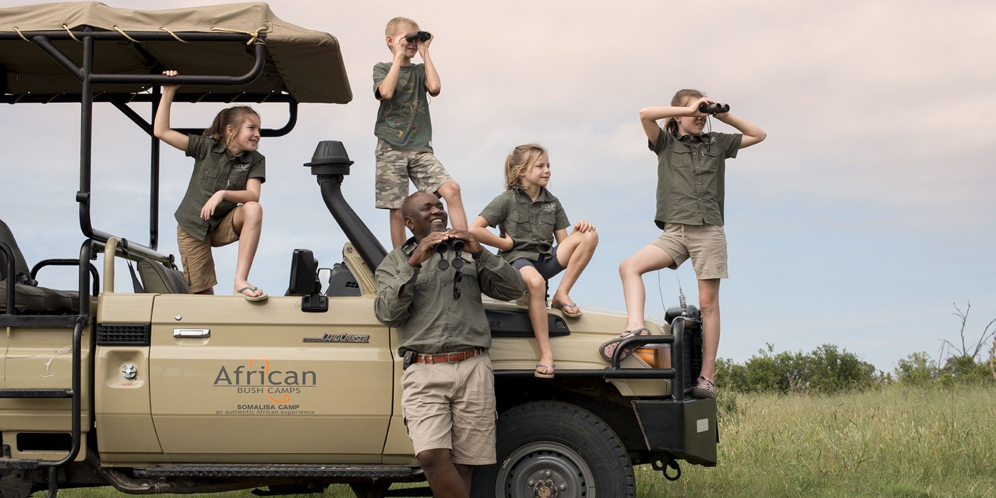 Four children with a guide on safari looking at wildlife through binoculars