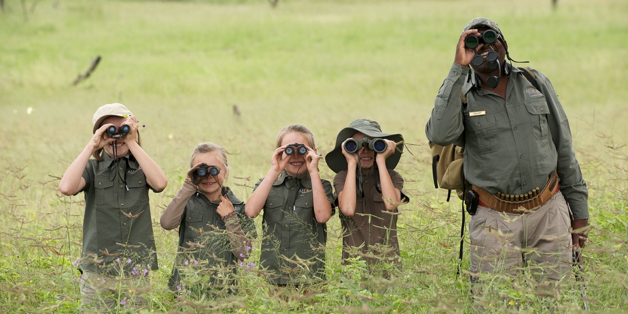 Four children with a guide looking through binoculars, standing in the long grass