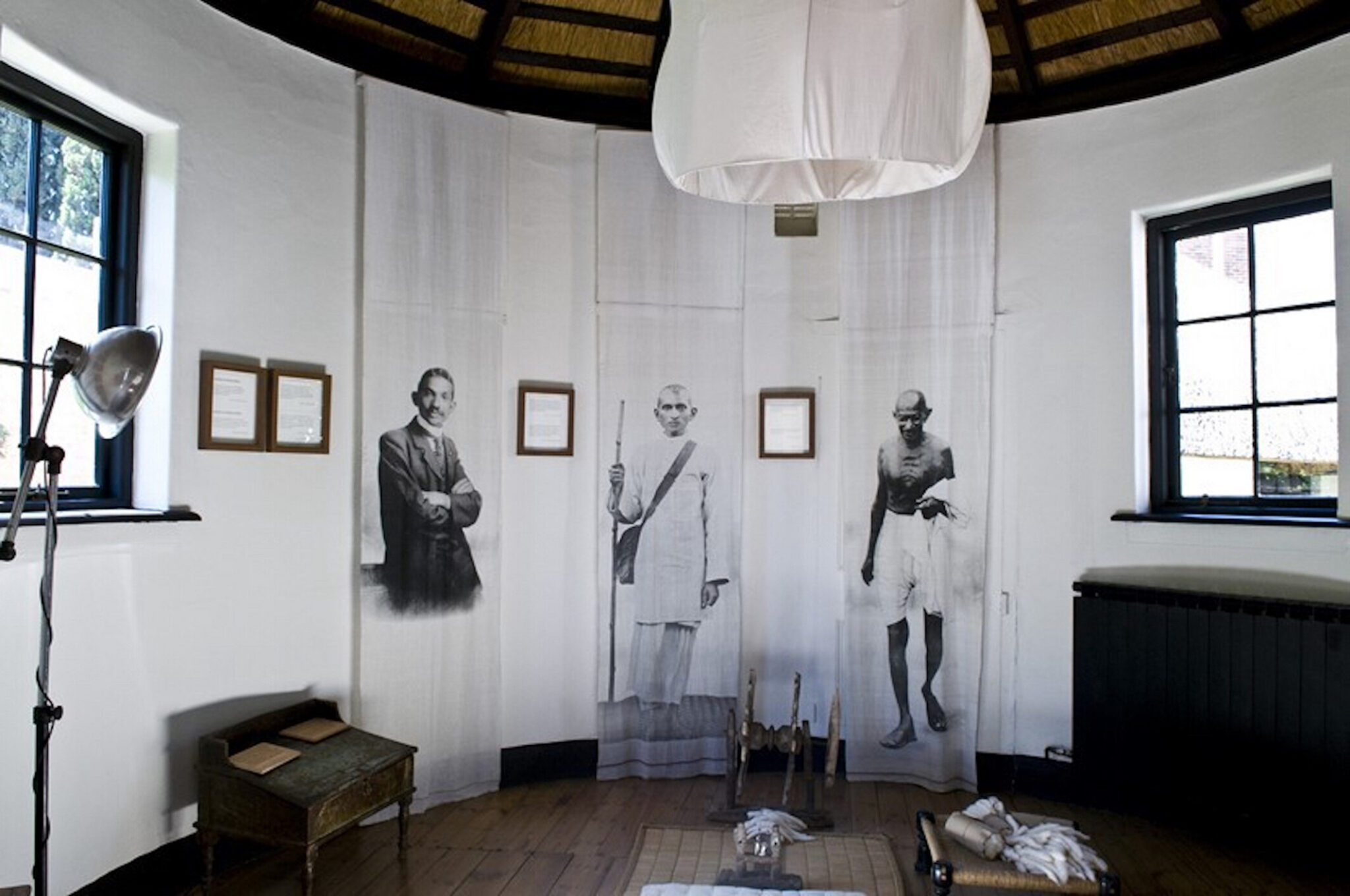 Interior of Satyagraha House featuring an exhibition room with large black-and-white portraits displayed on sheer panels, historic artifacts on the floor, and natural light from side windows.